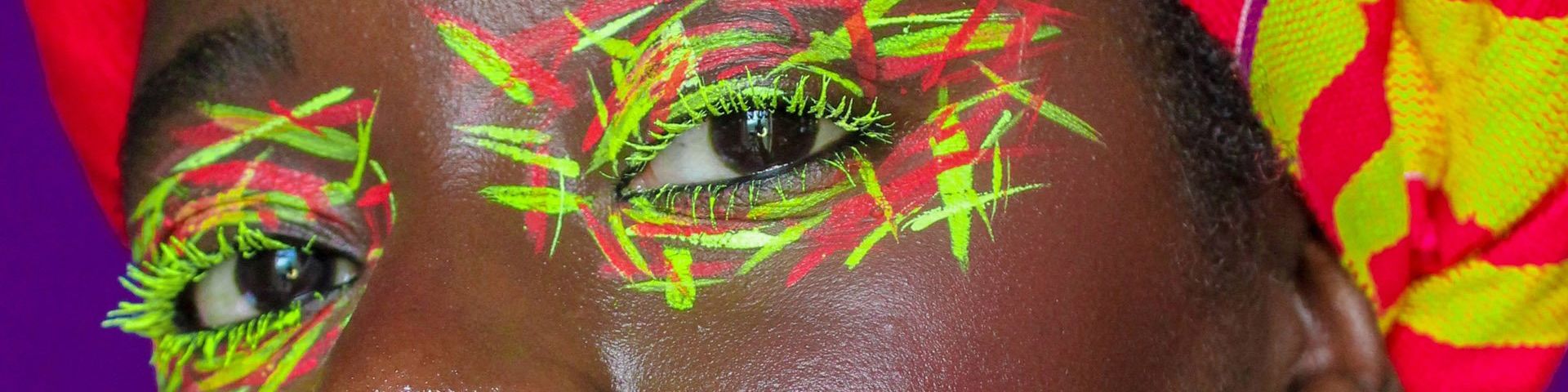 A woman’s eyes, painted with a neon yellow and red crosshatch pattern. She wears a matching head-wrap, which you can see just in shot. © Wendy Asumadu